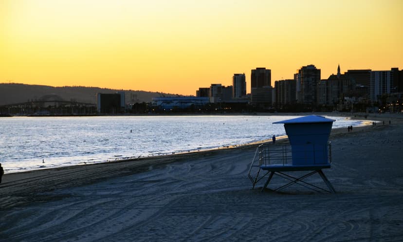 A lifeguard tower on the sand at sunset, with calm water and the Downtown Long Beach skyline in silhouette under a golden sky
