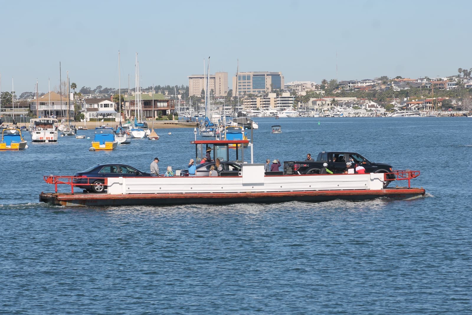 A small open ferry carries three cars and a dozen passengers across blue harbor water, with moored boats, homes and office towers behind
