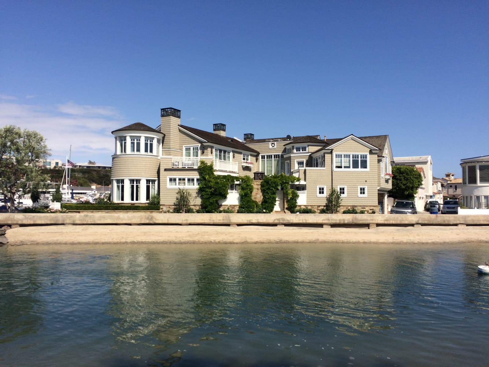 A large tan shingled house with a round corner tower and climbing vines stands behind a concrete seawall, a strip of sand and calm water