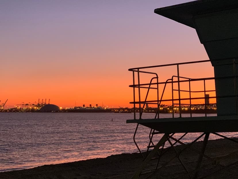 Silhouette of a lifeguard tower on the sand against an orange sky, with calm water and the lights of the port on the horizon