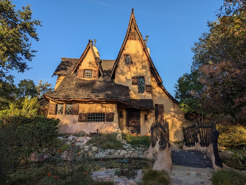 A storybook house with steep crooked shingle roofs and a wooden footbridge over a small pond, lit by low evening sun under a blue sky