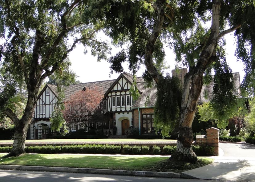 Tudor Revival house with white and dark timber gables, a wide front lawn and a low brick wall, framed by two large trees