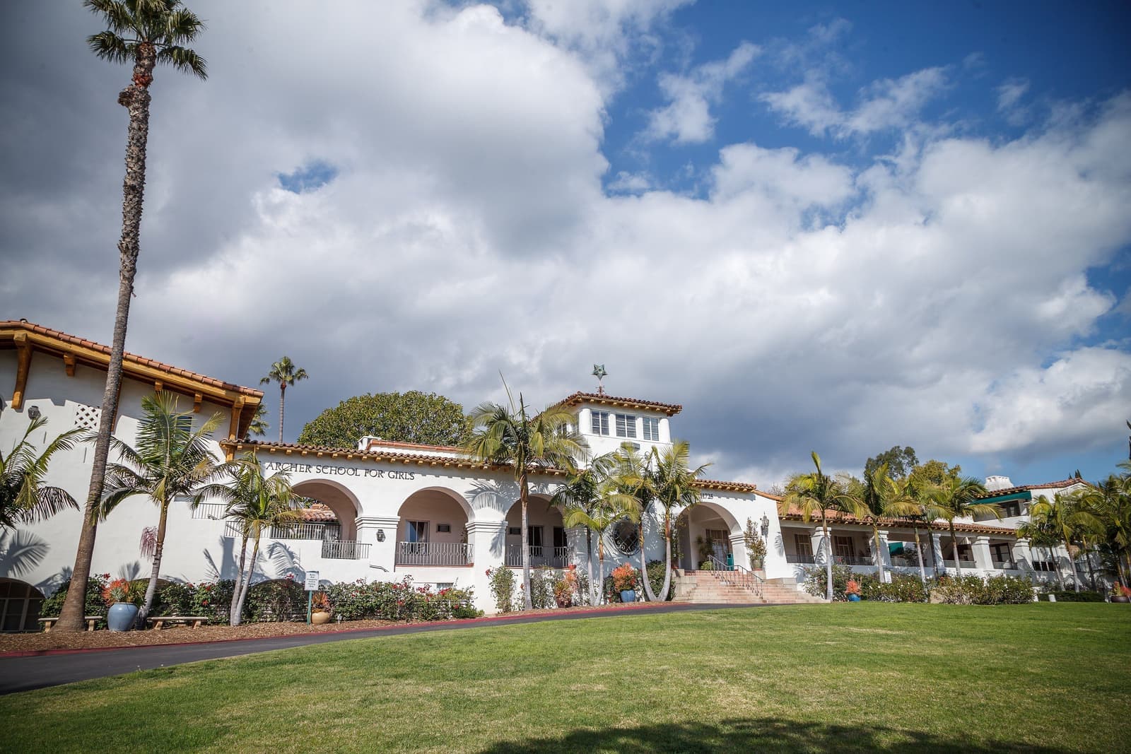 A long white Spanish Colonial building with arched arcades, a tower and red tile roofs behind a sloping lawn, palms and white clouds