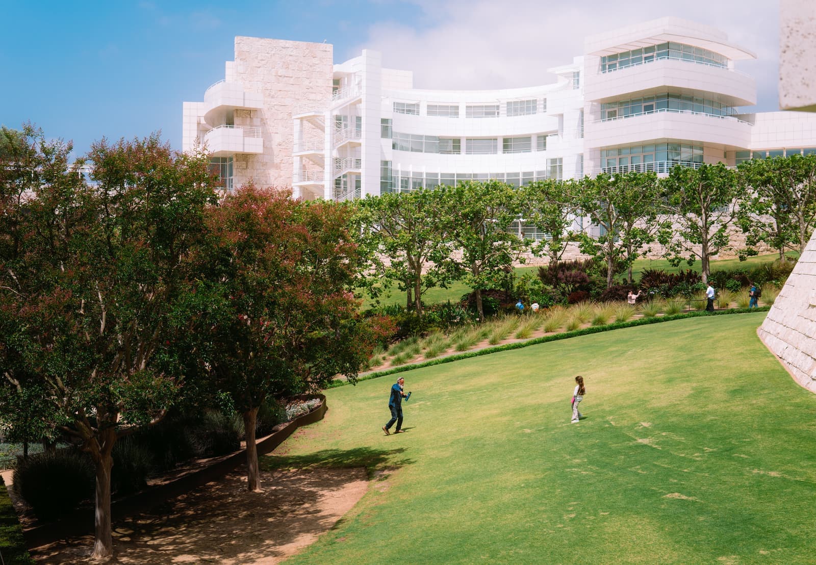 Curved white buildings of the Getty Center rising above a sloping green lawn and trees, with a few visitors on the grass