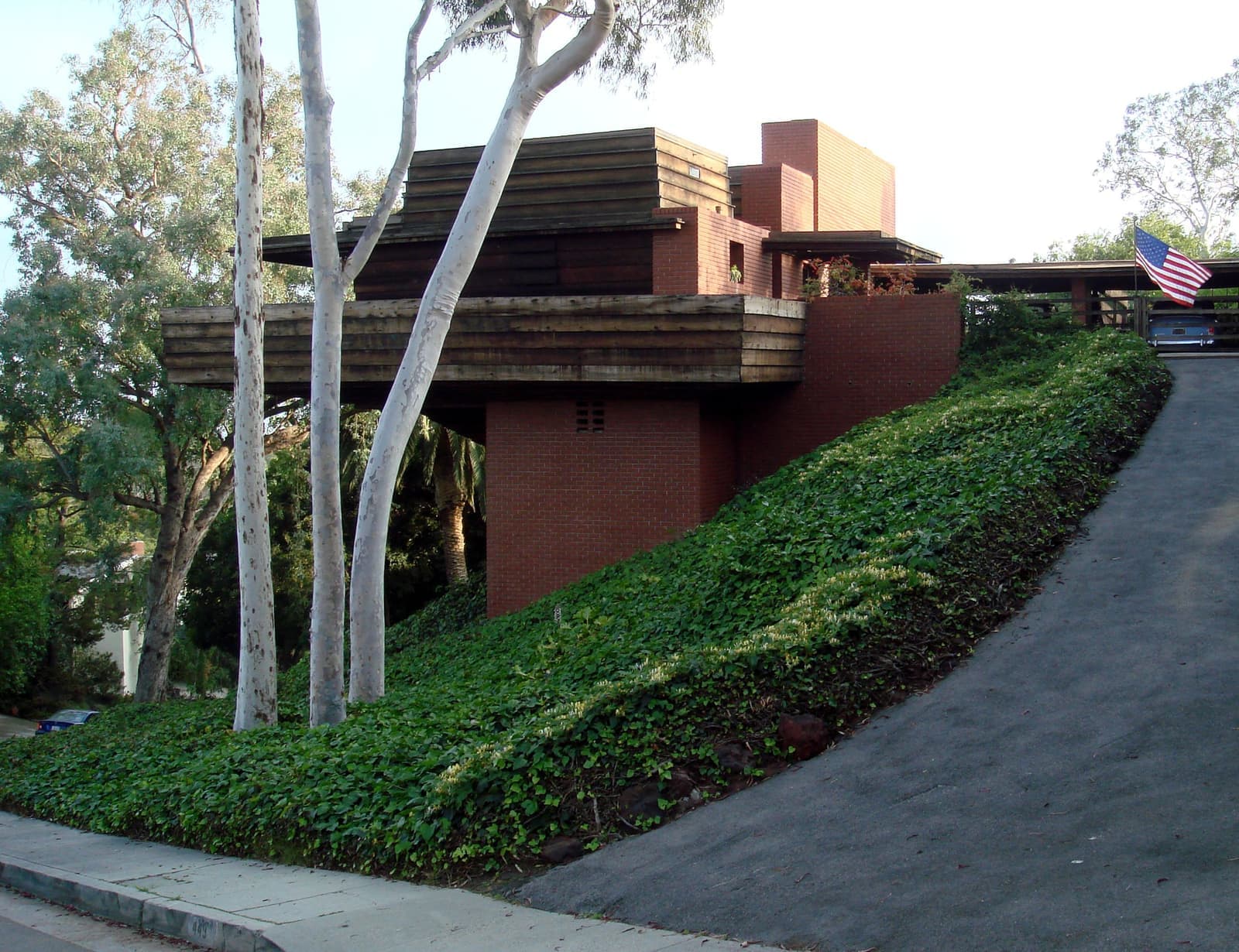 A brick and redwood house projecting over an ivy-covered slope beside a steep driveway, framed by pale eucalyptus trunks