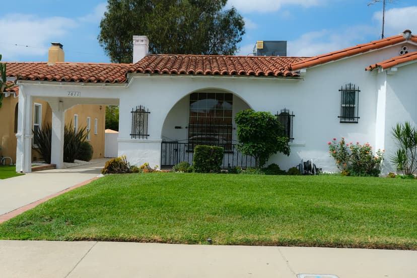 White stucco Spanish Revival bungalow with a red tile roof, an arched front window, iron window grilles and a green front lawn