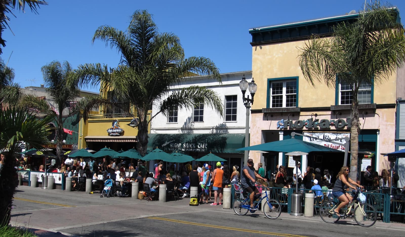 Busy sidewalk cafe under green umbrellas and palms, two people riding beach cruisers past two story storefronts