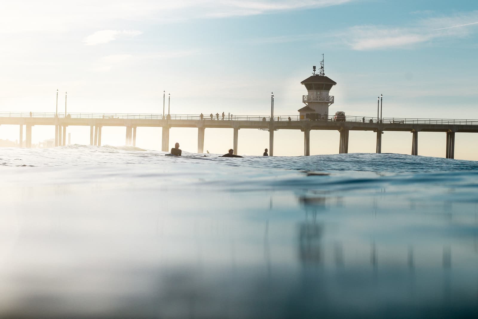 View from water level of the long pier and its lifeguard tower in soft hazy light, a few surfers sitting on their boards in the swell