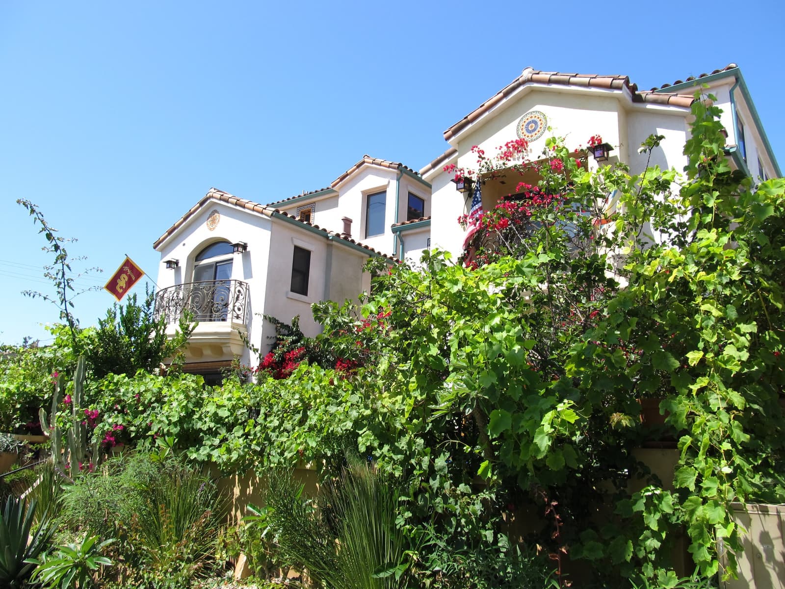 White stucco house with red tile roofs, arched windows and iron balconies, half hidden behind green vines and red bougainvillea
