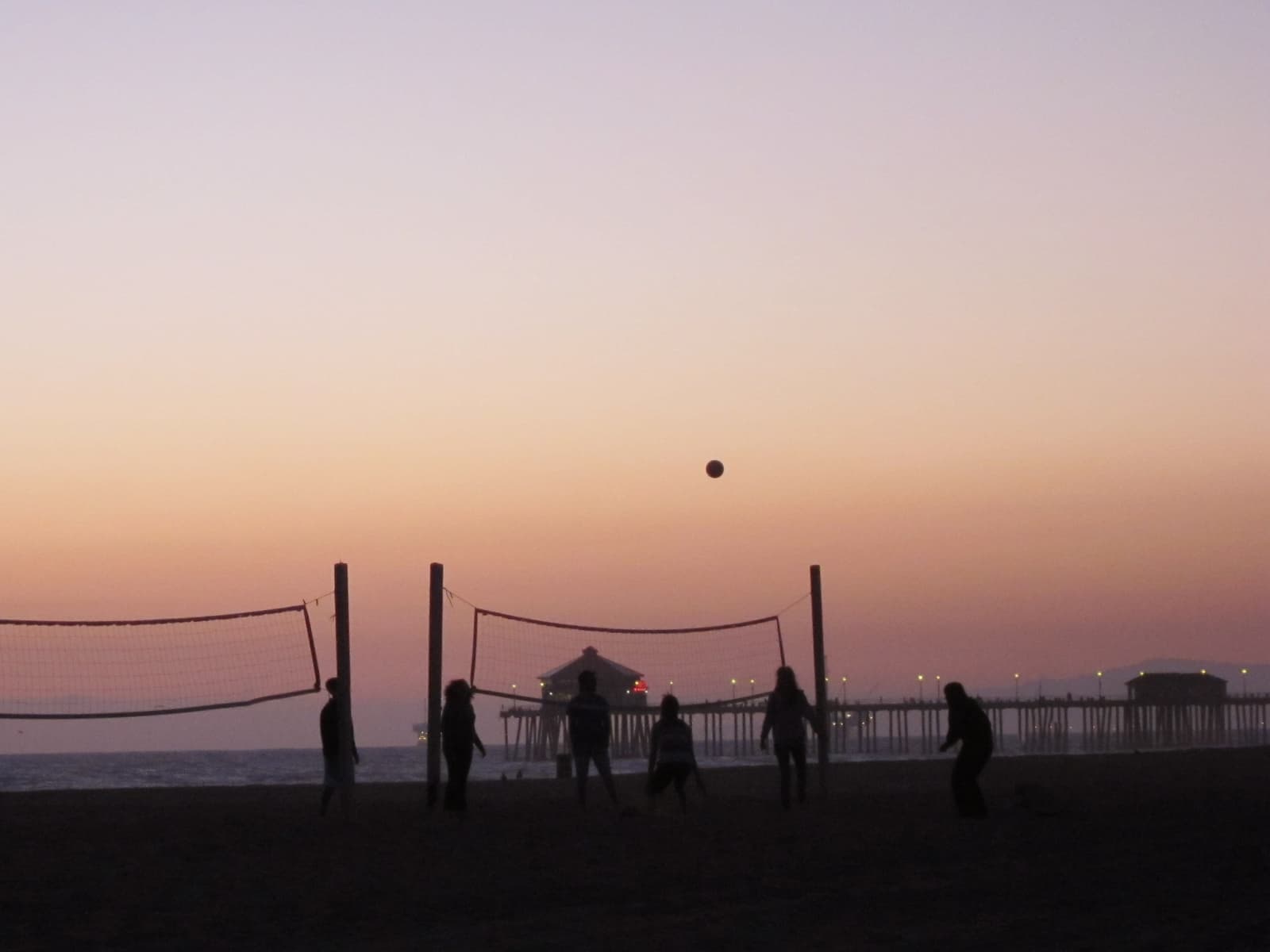 Silhouetted players at volleyball nets on the beach at dusk, a ball in midair, pink sky and the lit pier on the horizon