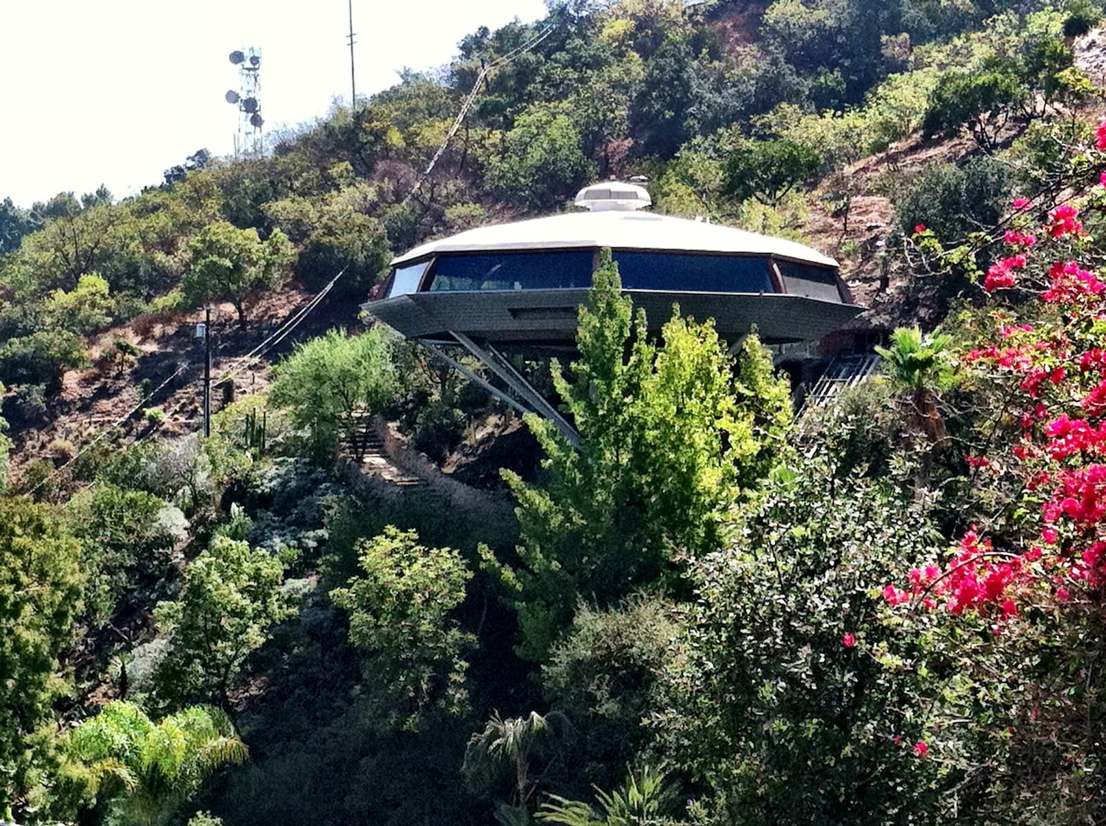 Octagonal house shaped like a flying saucer on a concrete pillar, on a steep brushy slope with pink bougainvillea in front
