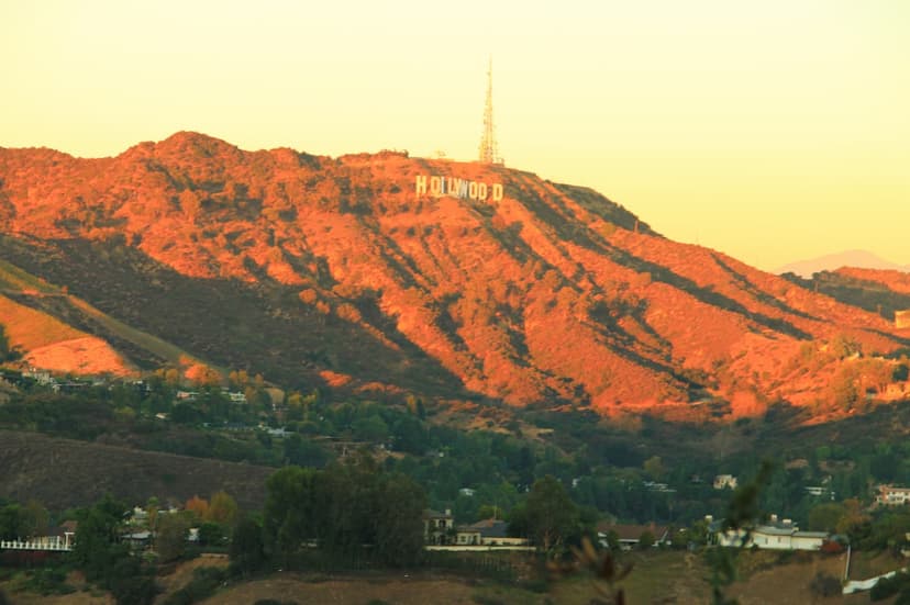 Mountain ridge glowing orange at sunset with the Hollywood Sign and a radio tower on top, green hills with homes below