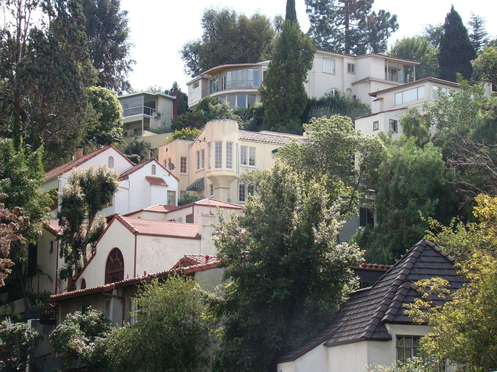 Cream and white stucco houses with red and gray tile roofs climbing a steep, tree-covered hillside, one above another