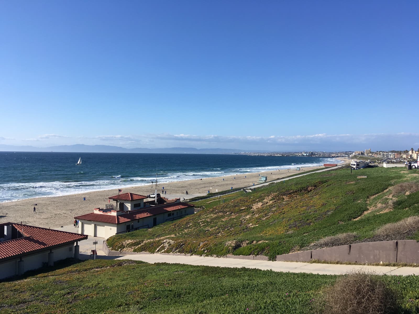 A green bluff covered in ice plant slopes down to Torrance Beach, with red tile beach buildings, a sailboat and the coast curving north