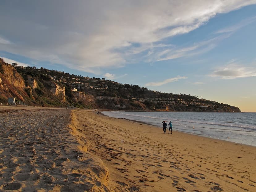 Two people walk along the wet sand of a wide beach in warm evening light, with tall brown cliffs and hillside homes in the distance
