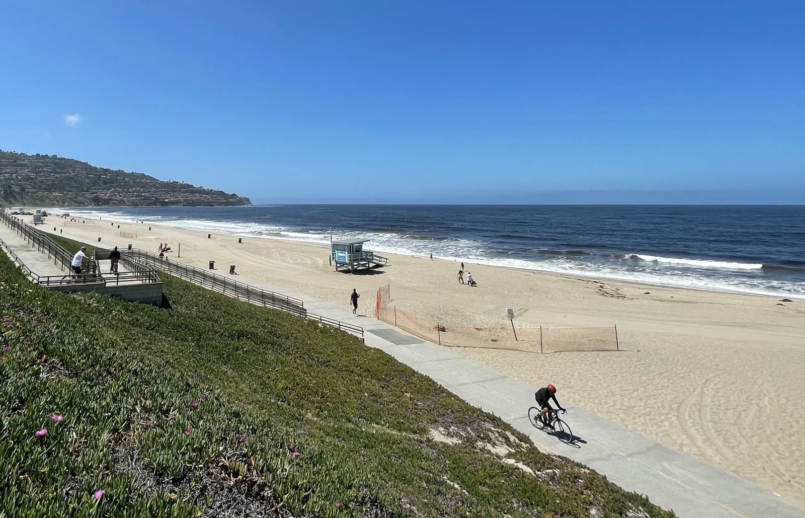A cyclist rides the paved path along Torrance Beach below a railing and green bluff, with the ocean and the Palos Verdes hill ahead