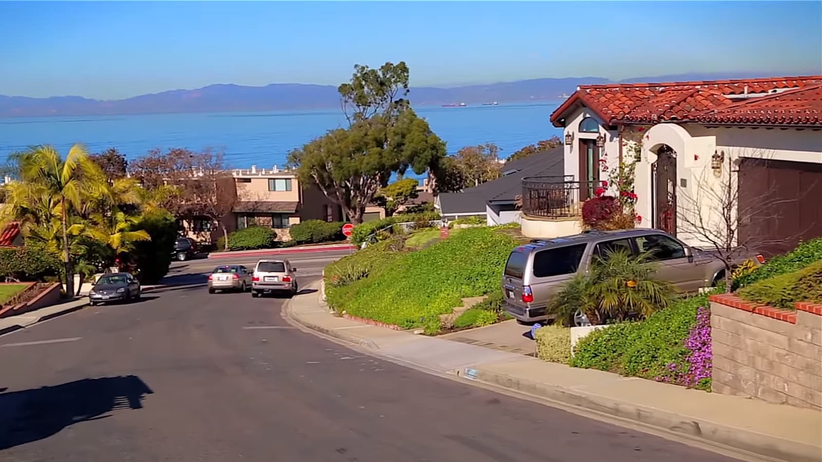 A curving street in the Hollywood Riviera with a white Spanish-style house, red tile roof and flowers, and blue ocean and mountains beyond