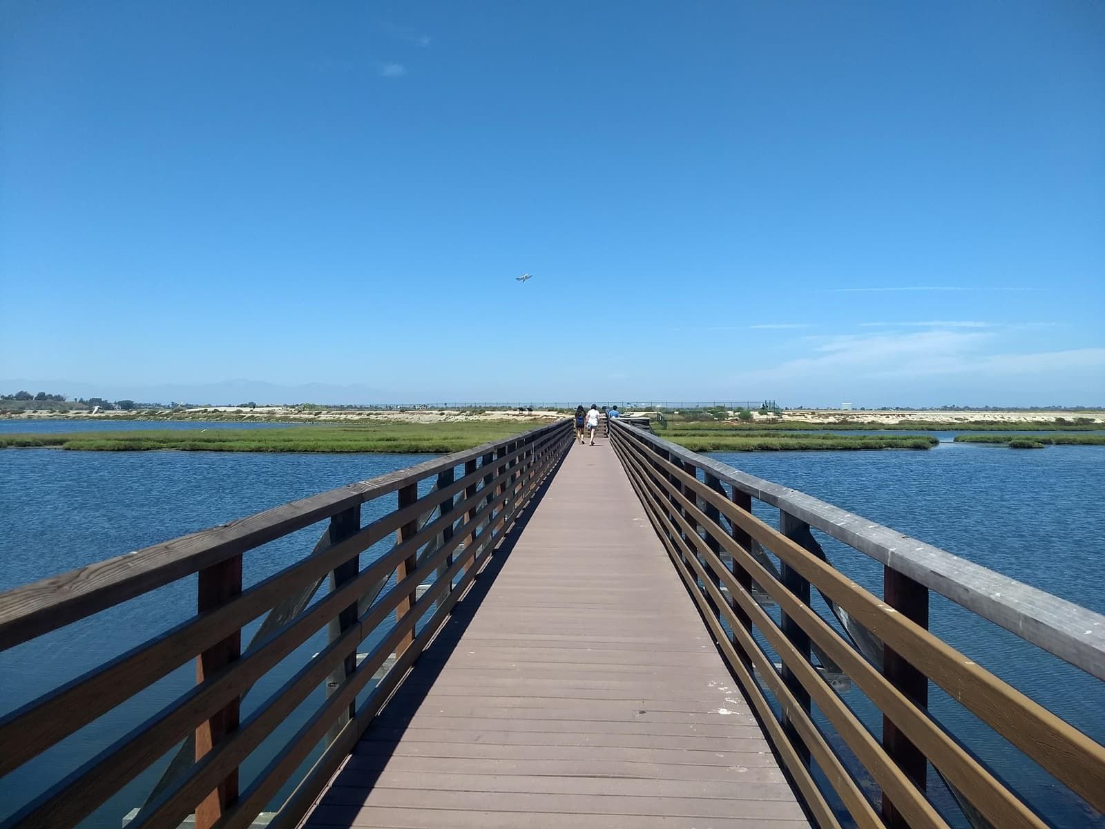 Long wooden footbridge with railings running straight out over blue water and green marsh, a few walkers far down it under a clear sky
