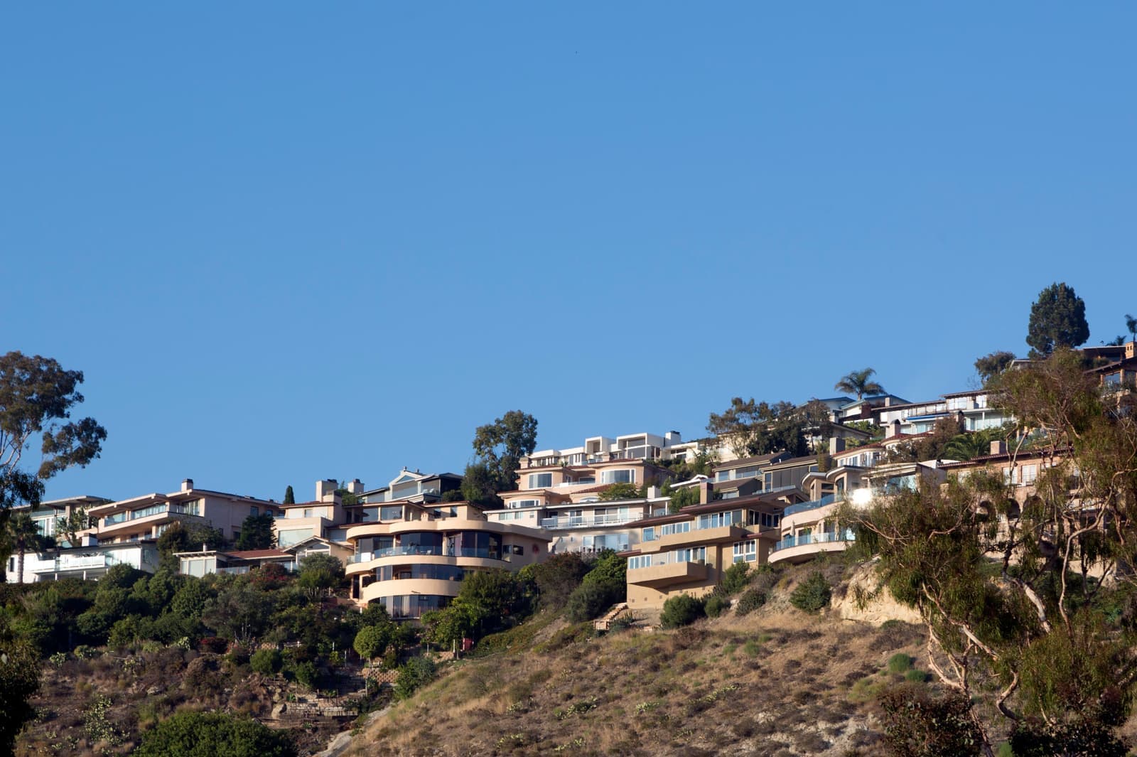 Tiered hillside homes with balconies above a slope of dry brush and trees, under a clear blue sky