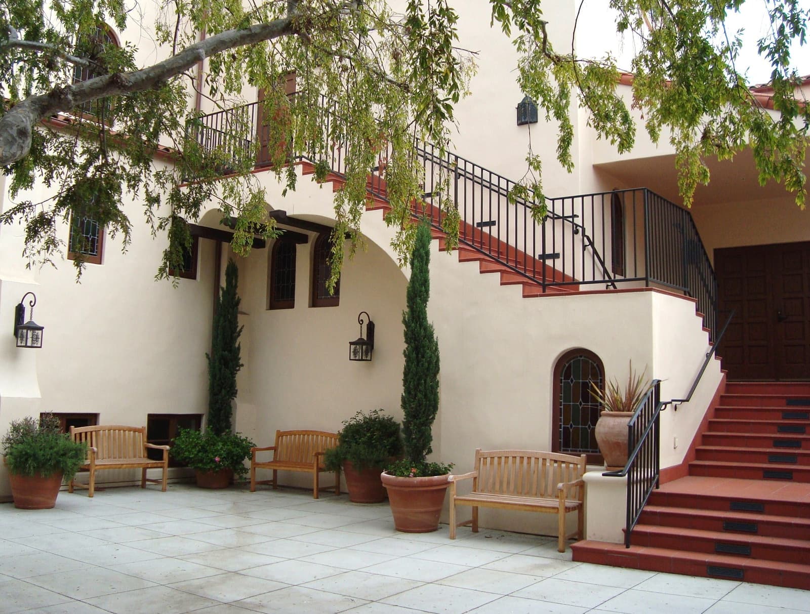 A white stucco courtyard with red tiled stairs, iron railings, arched windows, teak benches and potted cypress under a leafy branch
