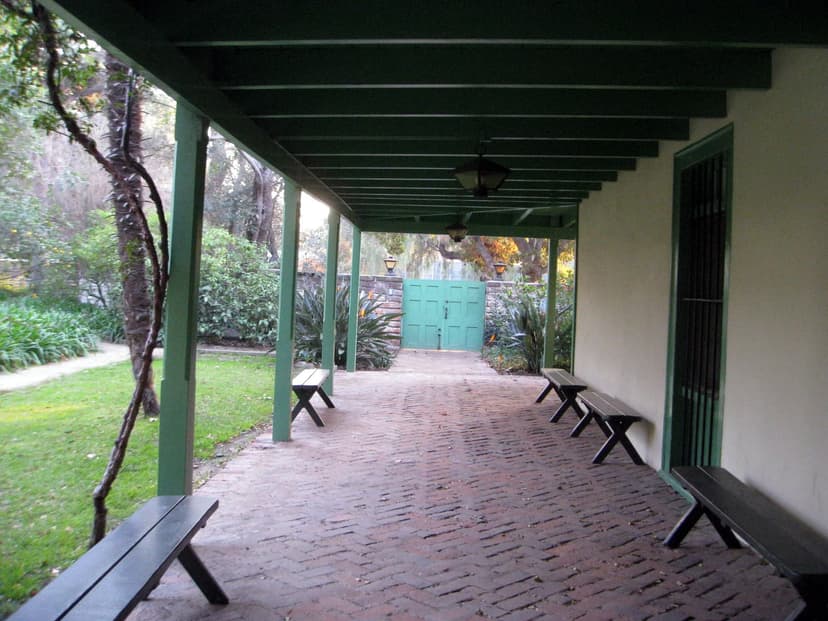 A shaded veranda with green wooden posts, a herringbone brick floor and dark benches leading to a green double gate in a stone wall