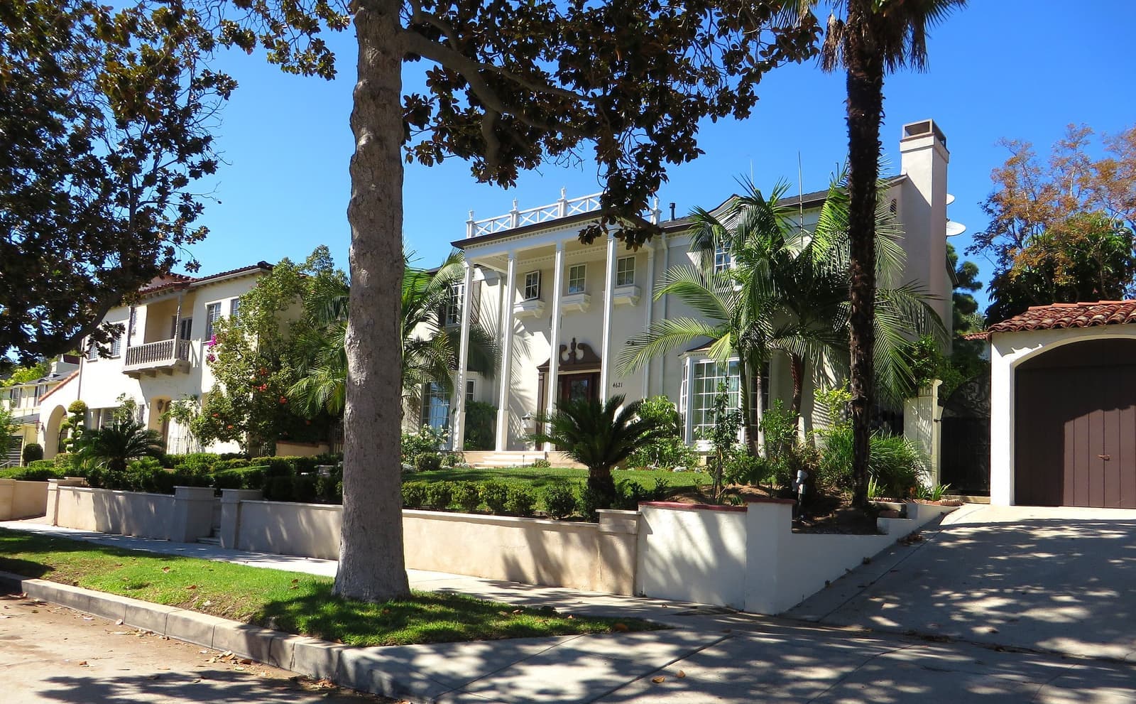 A white columned Colonial Revival house and a Spanish style house behind low garden walls, with a magnolia and palms on a sunny day