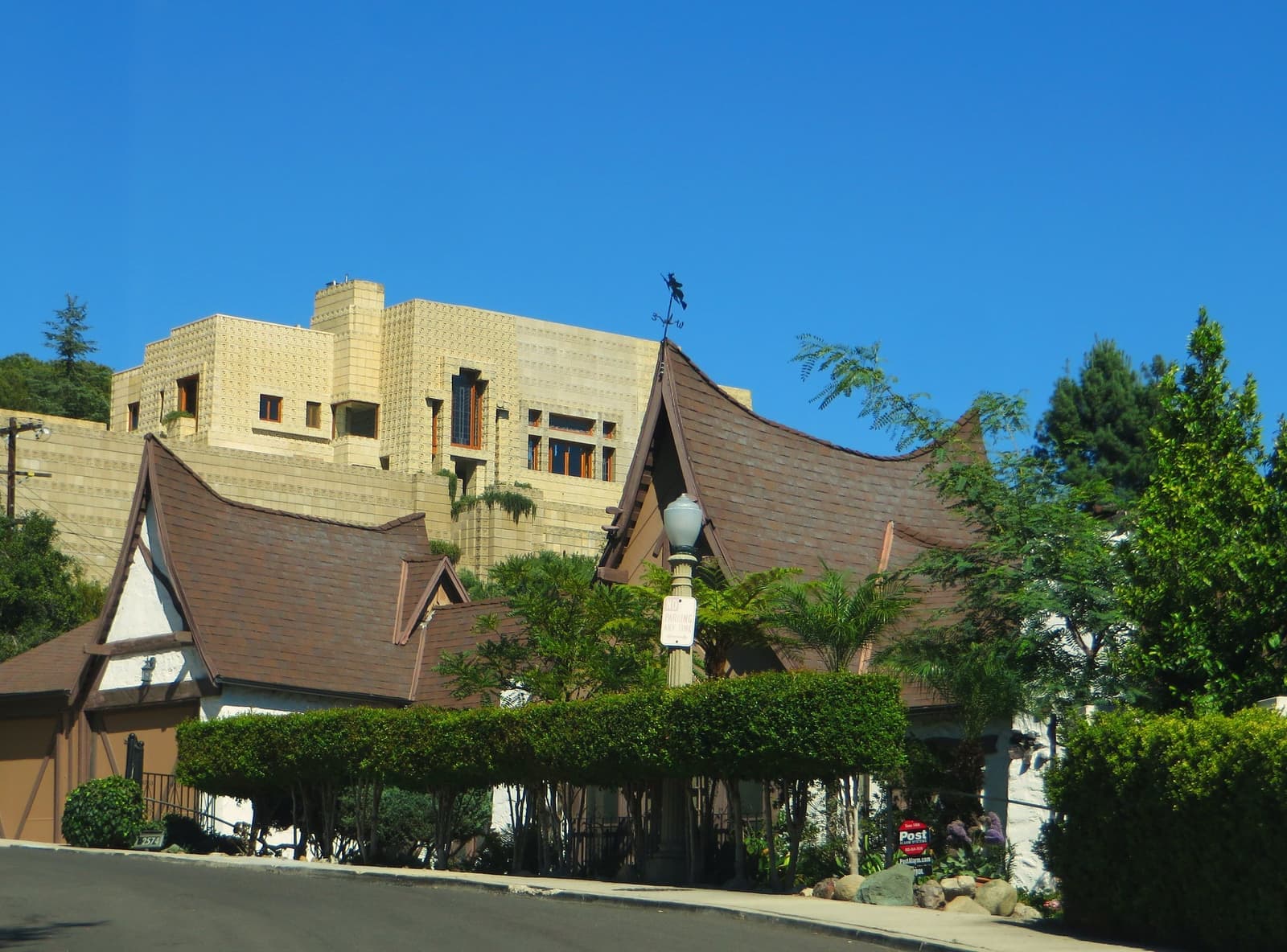 The swooping shingled roof of a fairy tale style house among green plants, with the patterned concrete blocks of the Ennis House above