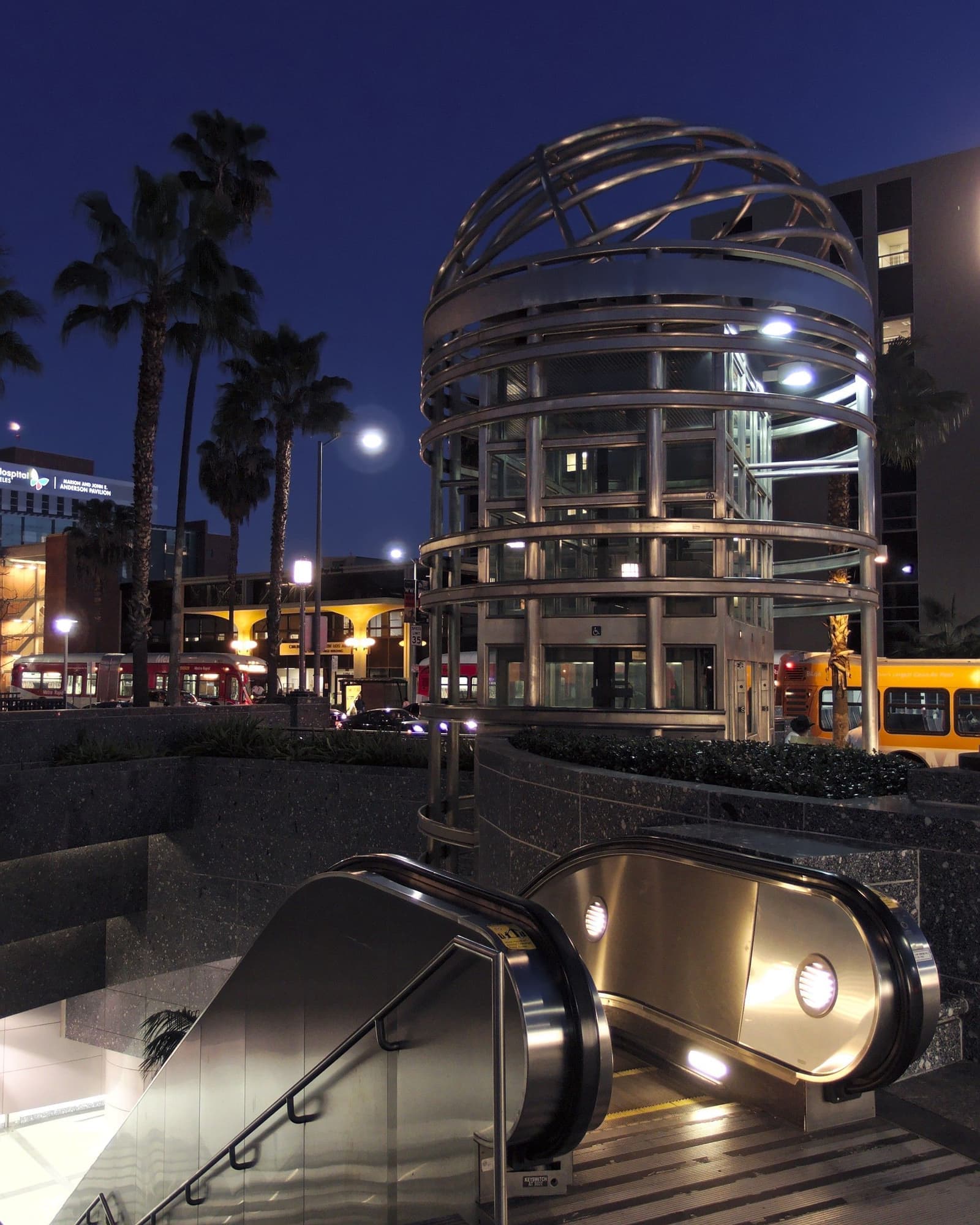 A round glass and steel elevator tower and escalators at a subway entrance under a deep blue evening sky, with palms and bus lights