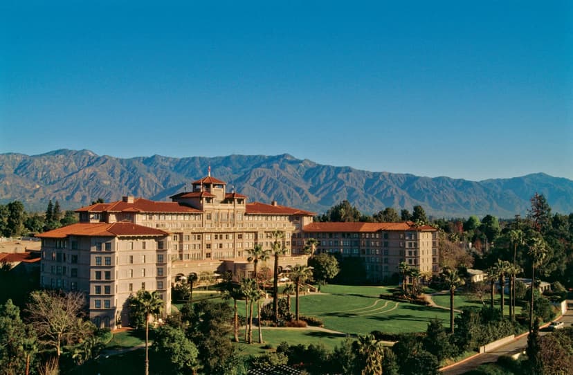 Large hotel with red tile roofs and a central tower above lawns and palm trees, with a long mountain range under a clear blue sky