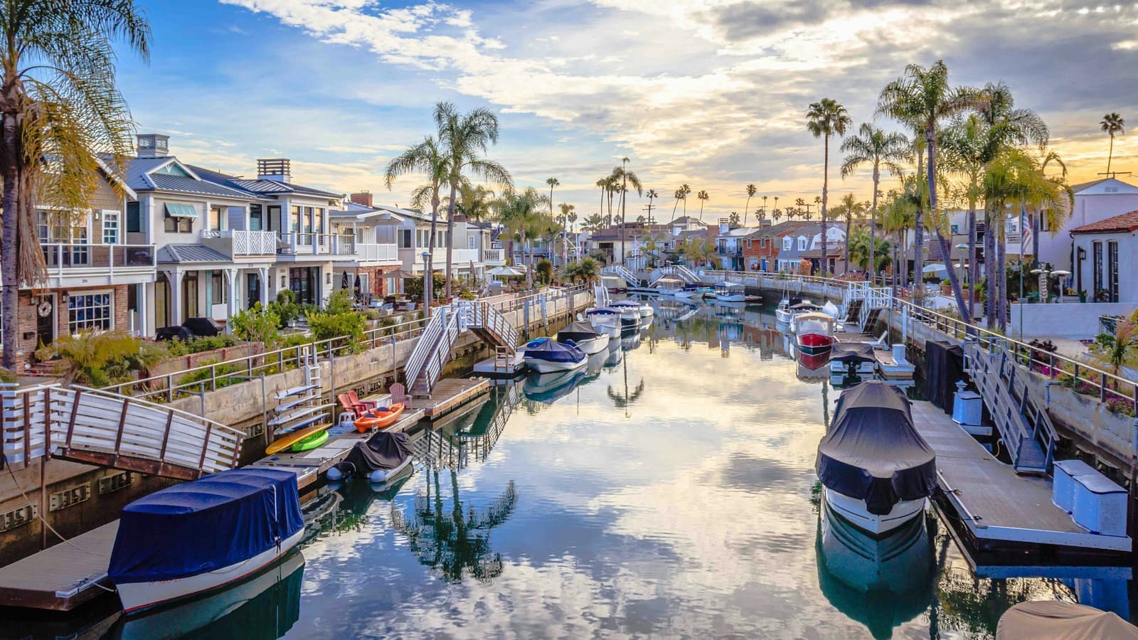 A long, calm canal in Naples with covered boats at private docks, palm trees and homes on both banks under a cloudy sky