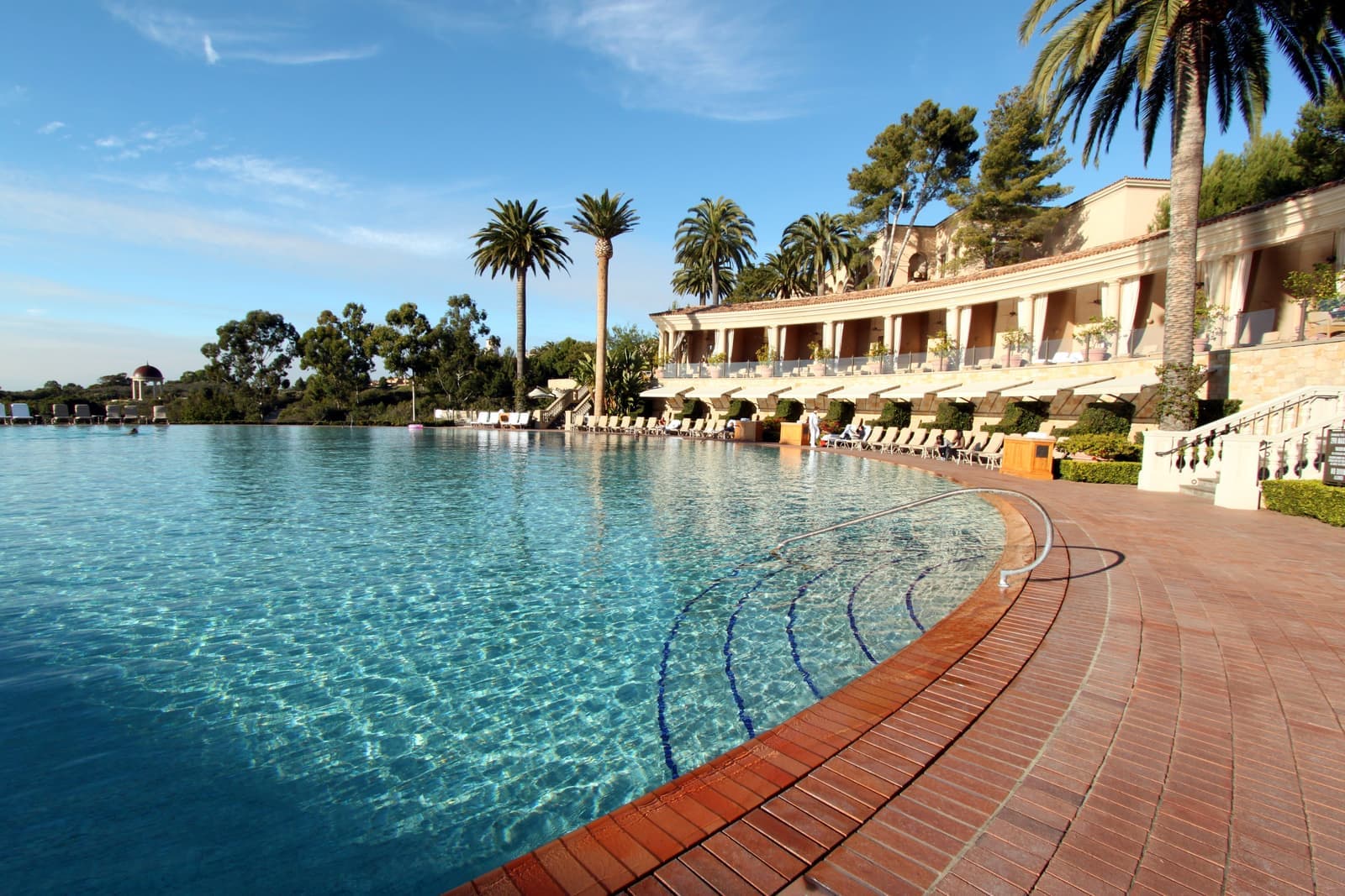 A large round pool with clear turquoise water, a curved brick deck, palm trees and a long cream colonnade under a blue sky