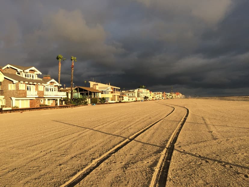 Shingled beach houses and two tall palms glow in golden light beside a wide, empty beach with tire tracks, under a dark gray sky