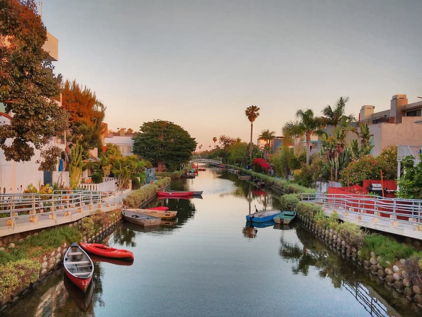 A Venice canal at sunset, with red and blue canoes on the water and a white footbridge in the distance