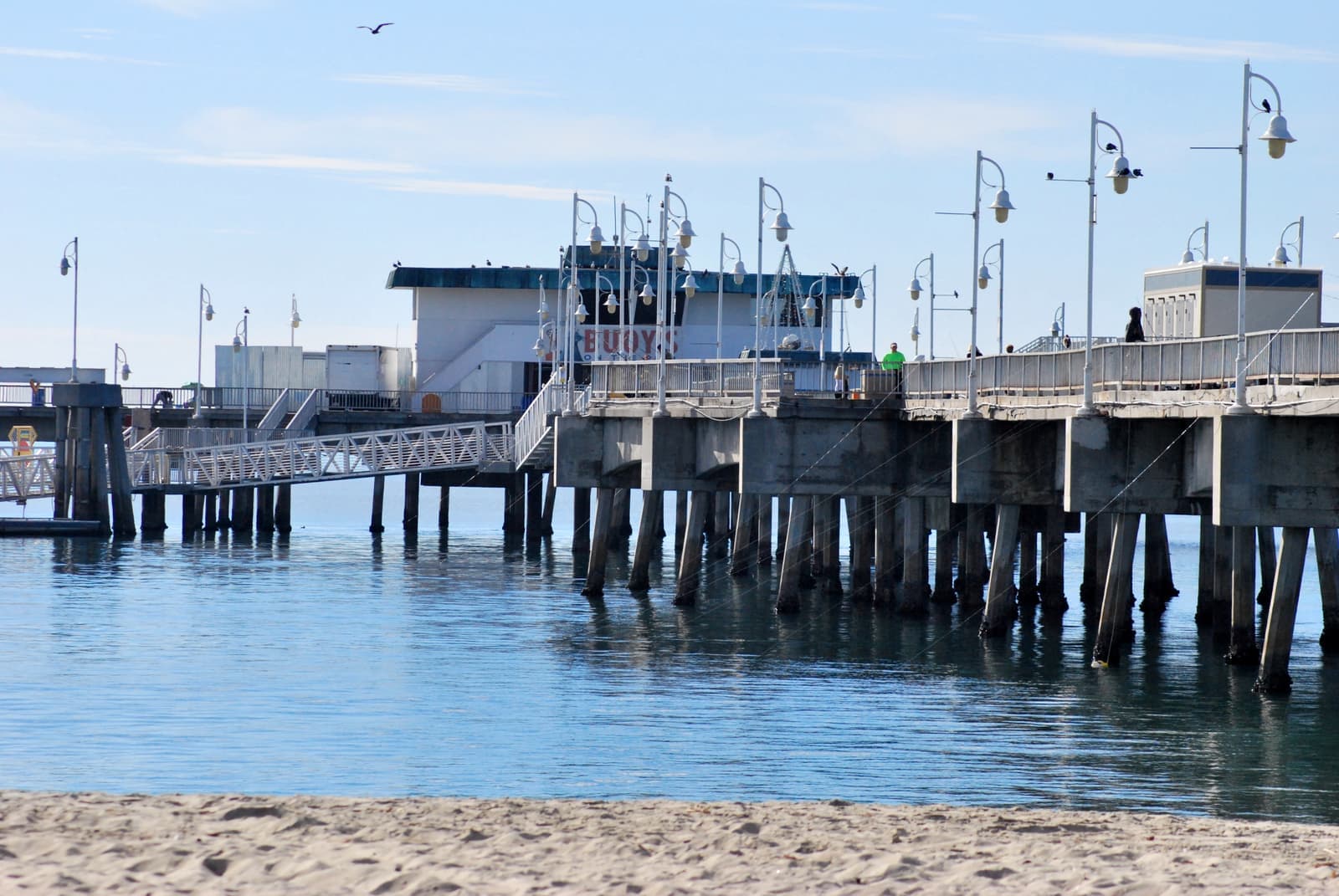 The concrete pilings and lamp posts of Belmont Veterans Memorial Pier over calm blue water, with sand in the foreground