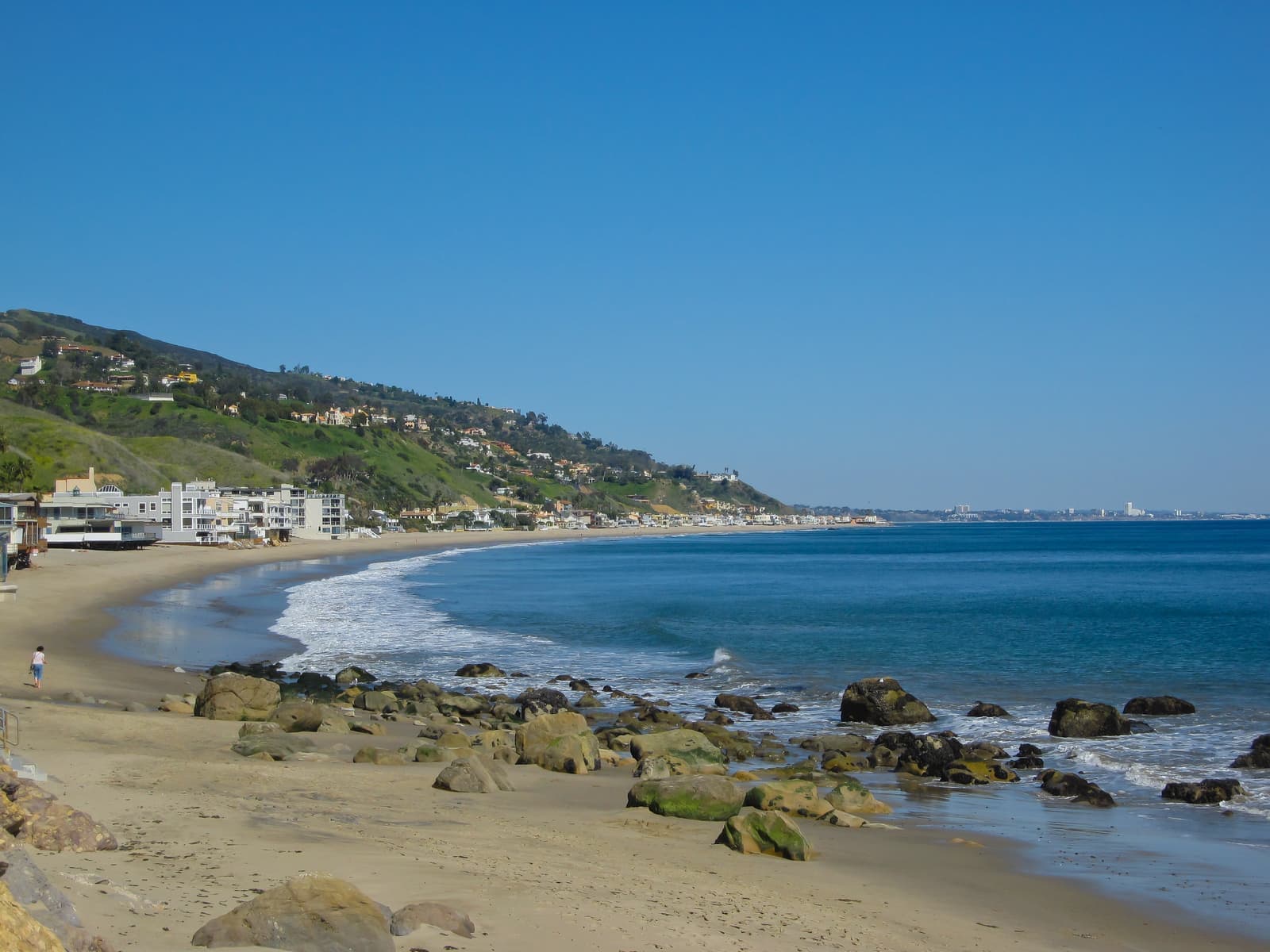 A sandy beach curving into the distance, rounded boulders at the waterline, white beachfront buildings at left and green hills above