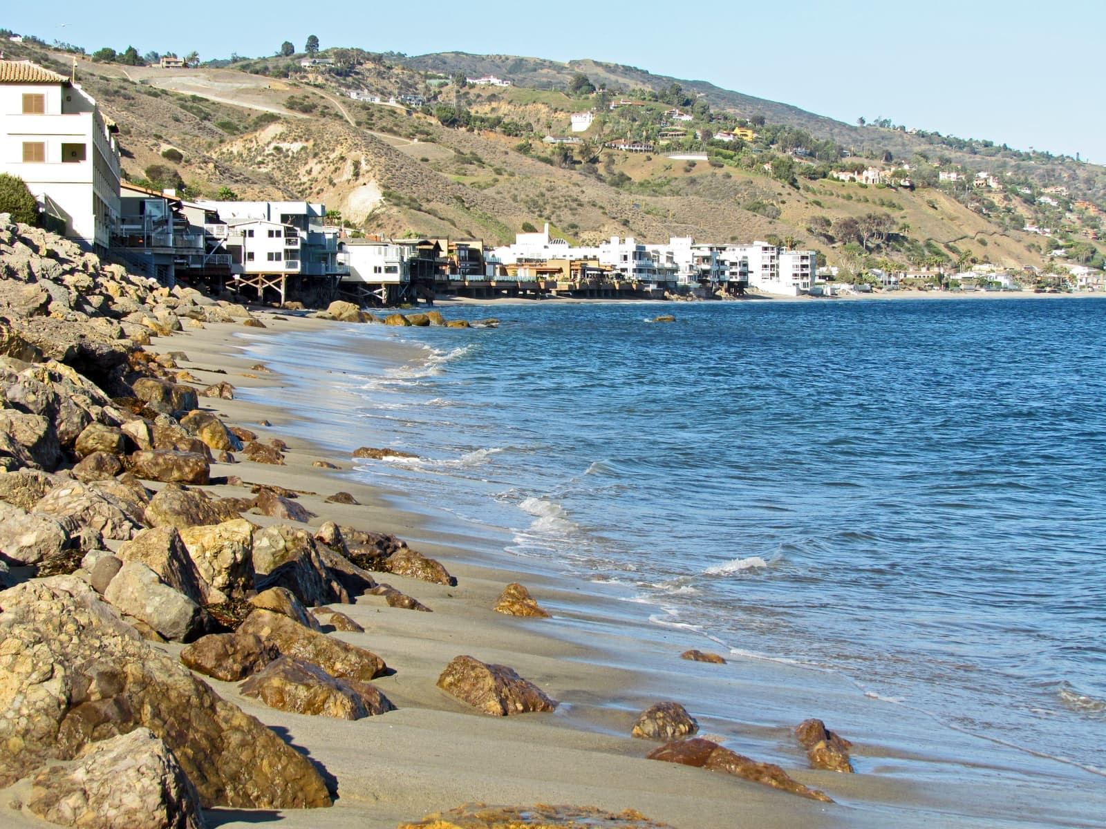 Boulders and wet sand in the foreground, calm blue water at right, and a row of beach houses on wooden pilings below dry hills