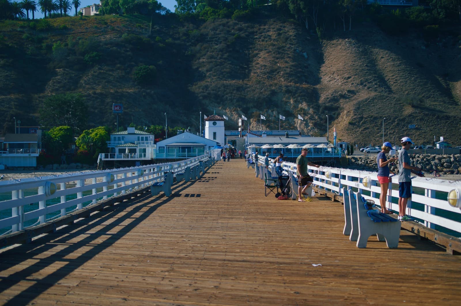 A long wooden pier deck with white railings and benches, people fishing on the right, white pier buildings and a brown hillside ahead
