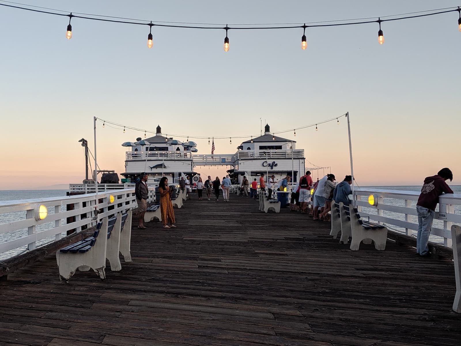 People stroll a wooden pier under strings of lights toward two white buildings at its end, under a pink evening sky