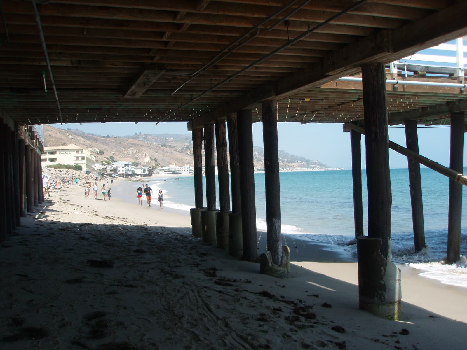 Seen from under the Malibu Pier, wooden pilings frame people walking along the water toward a hotel, beach houses and hills