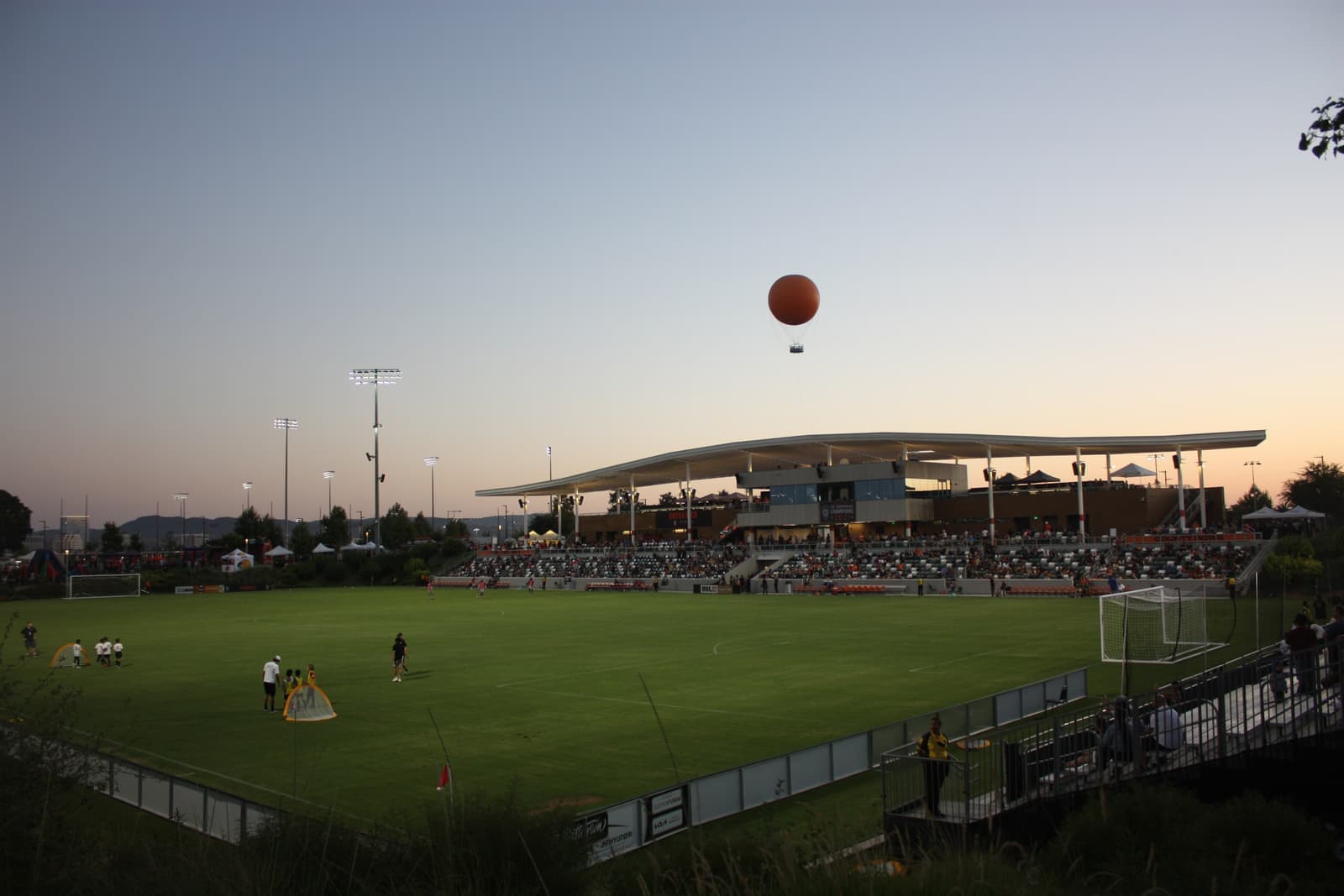 Soccer stadium at dusk with a covered grandstand full of fans, players warming up on the field and an orange balloon in the sky