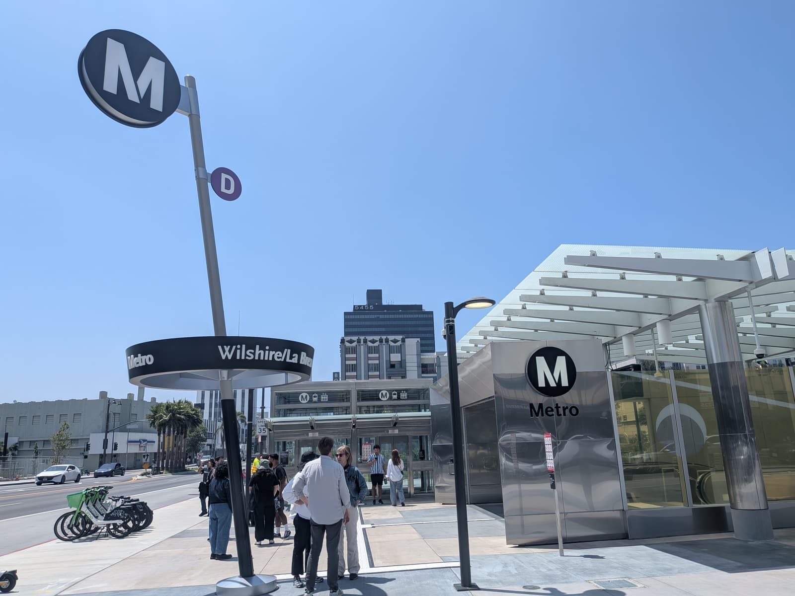 Вход в метро из нержавеющей стали со знаком Metro и указателем Wilshire/La Brea, люди на площади и велосипеды рядом