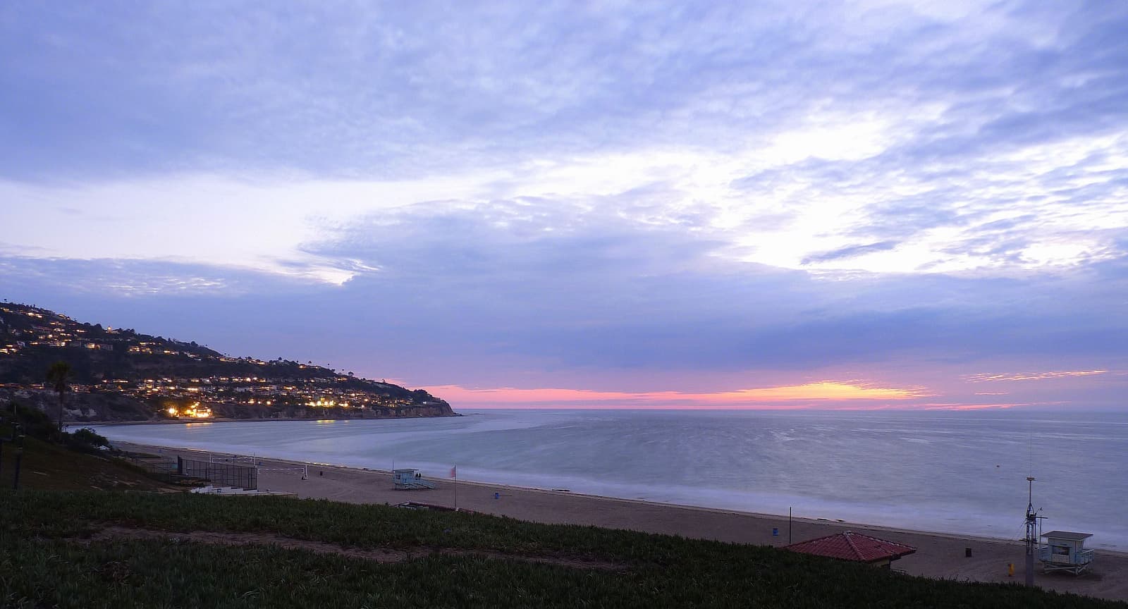 Dusk over a wide beach and calm ocean, a lifeguard tower on the sand, and house lights on the dark Palos Verdes hill to the left