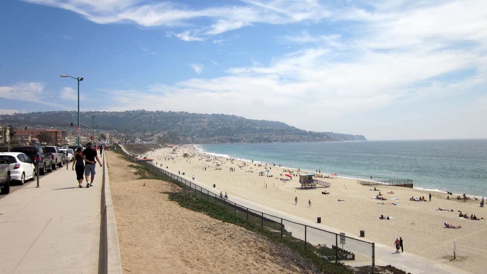 People walk along the Esplanade above a busy beach with a lifeguard tower, and the green Palos Verdes hill rises at the far end of the shore