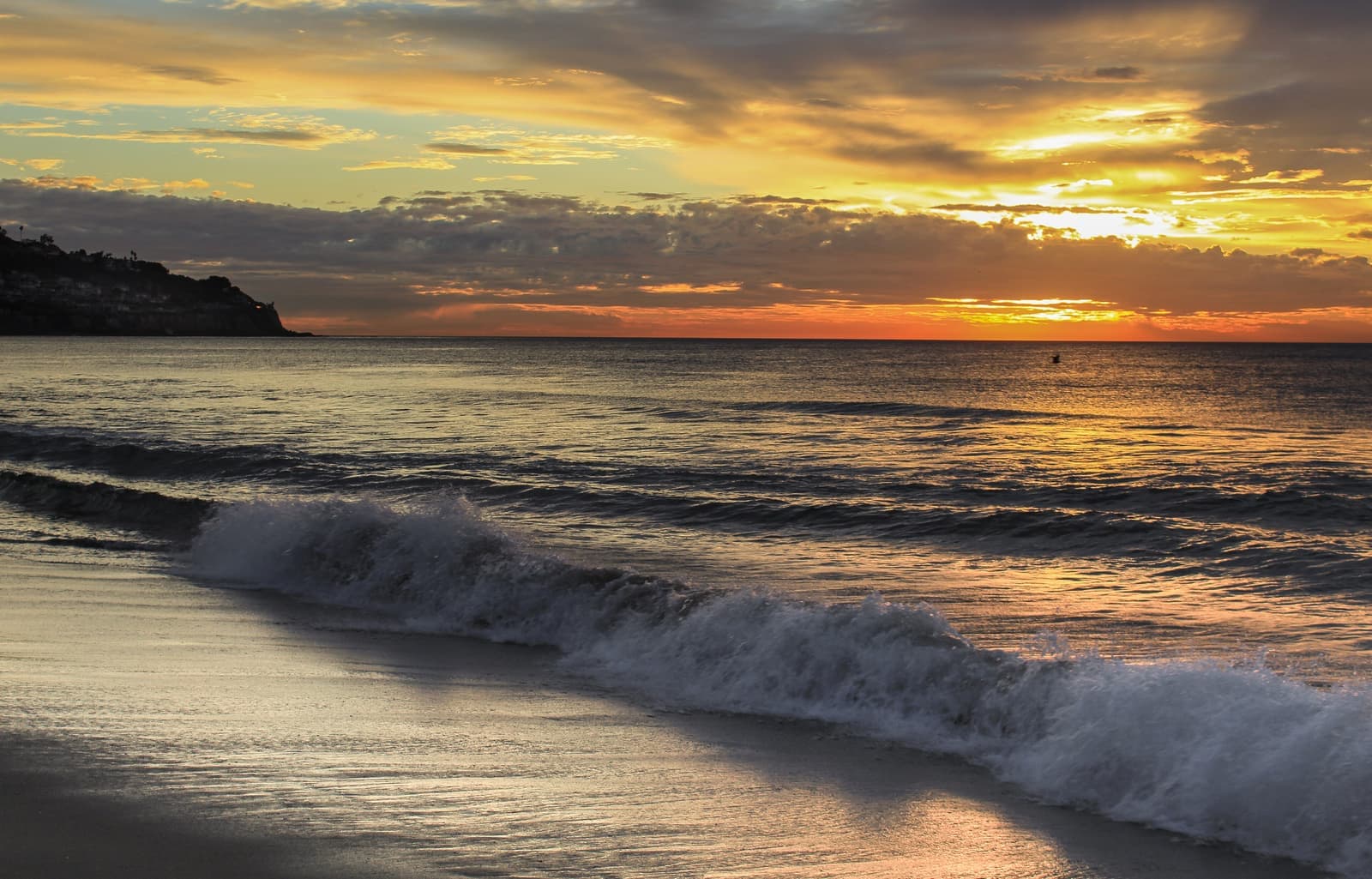A wave breaks on the sand under an orange and gold sunset sky, with a dark headland at the left end of the horizon