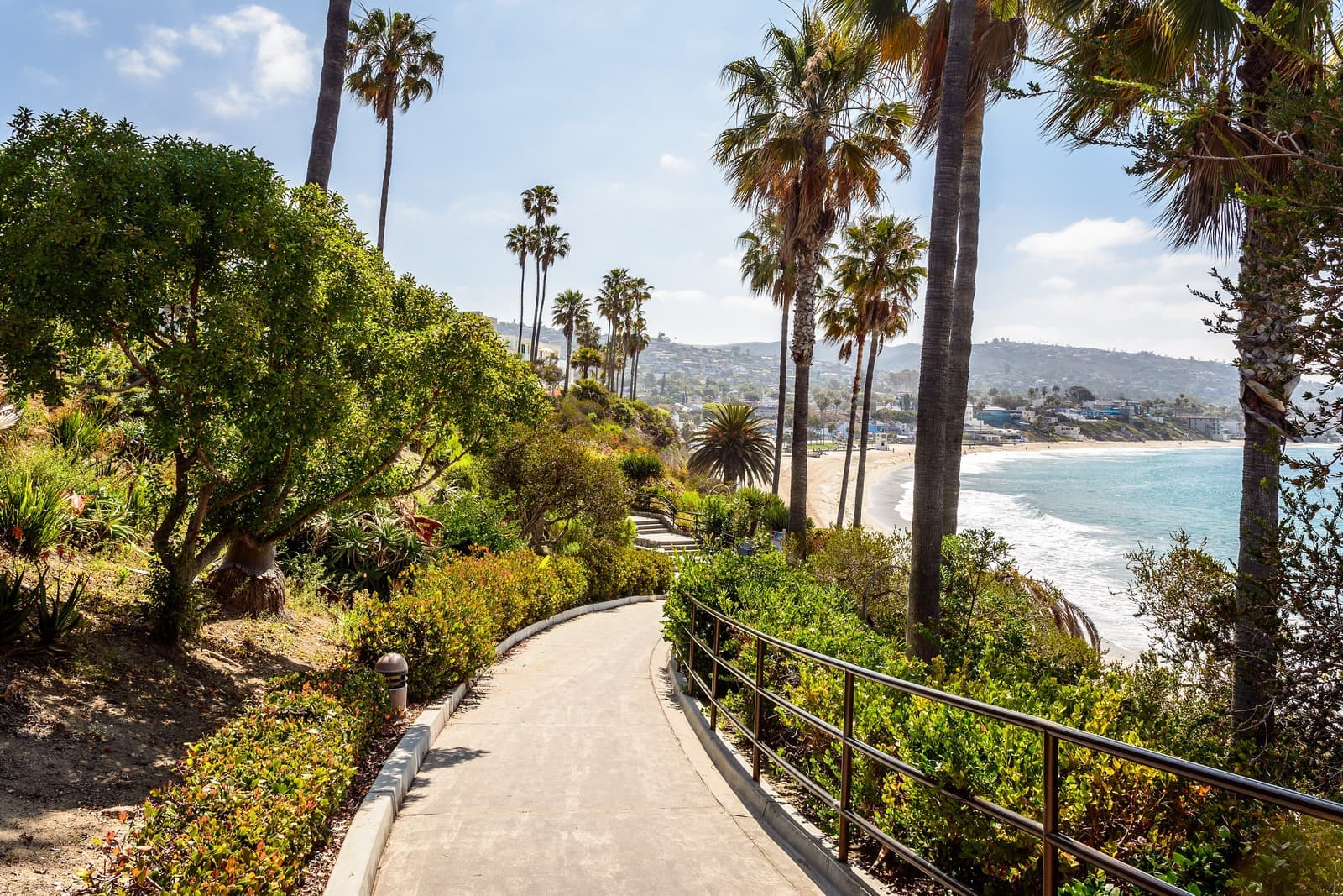 A paved path with a railing winds through gardens and tall palms on the bluff, a sandy beach, surf and hillside homes beyond