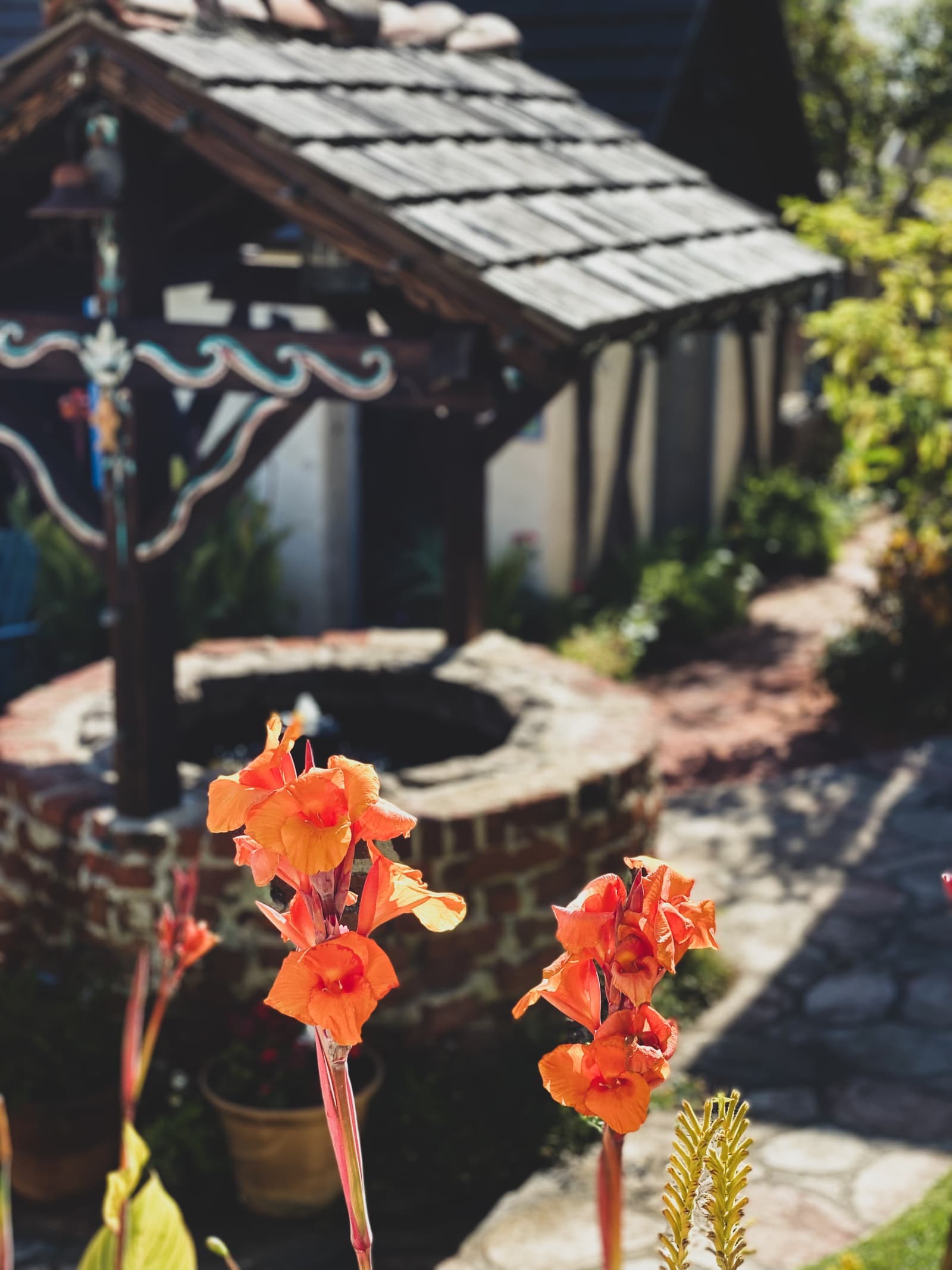 Orange canna lilies in front of a brick garden well with a shingled roof, a flagstone path and a small cottage behind