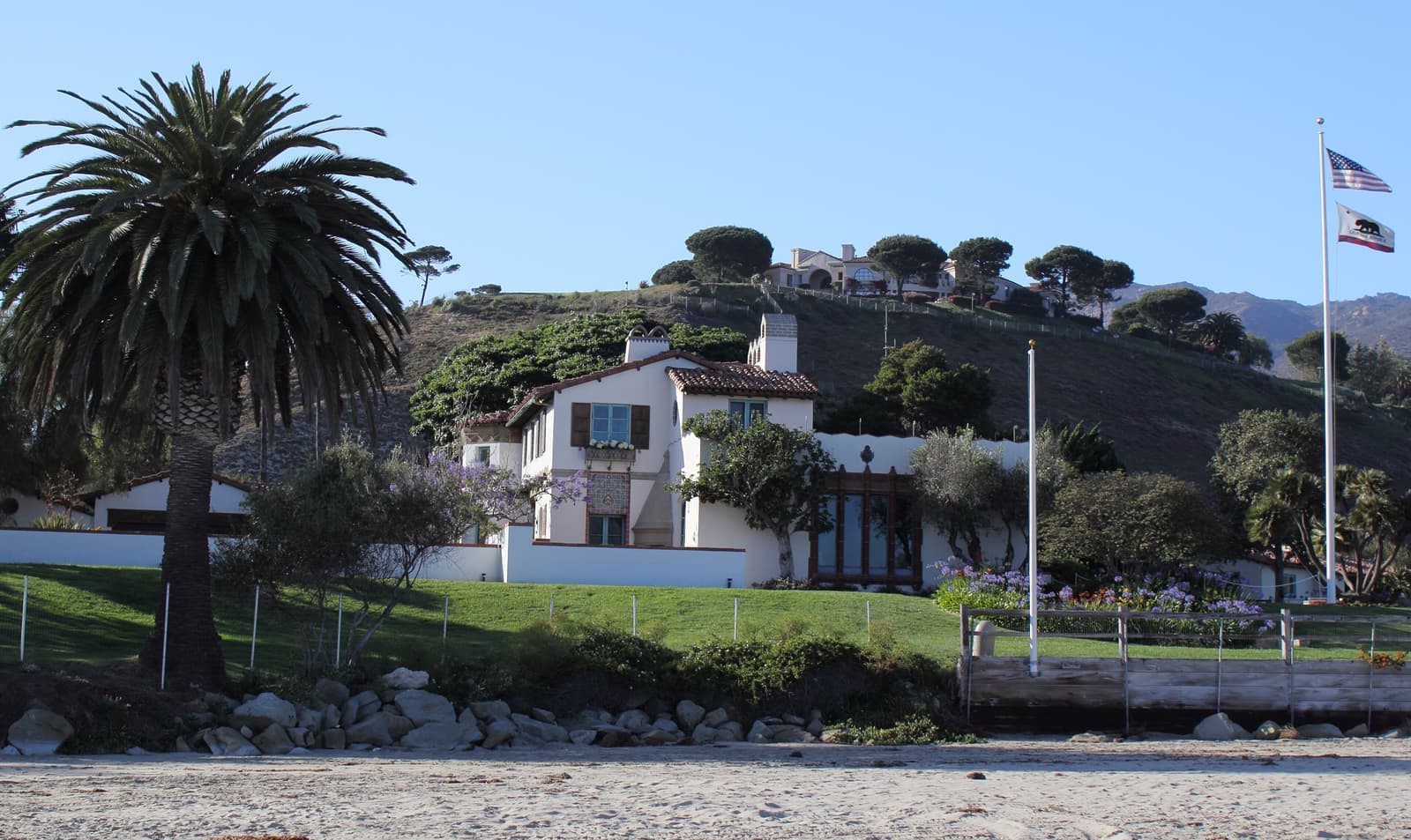 A white Spanish Colonial house with a red tile roof and a tall palm, on a lawn above a sandy beach, with a hillside behind