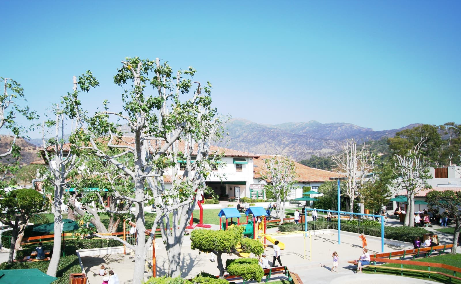 A sunny courtyard with pale pruned sycamores, a playground, lawn and low white shops with tile roofs, brown mountains behind