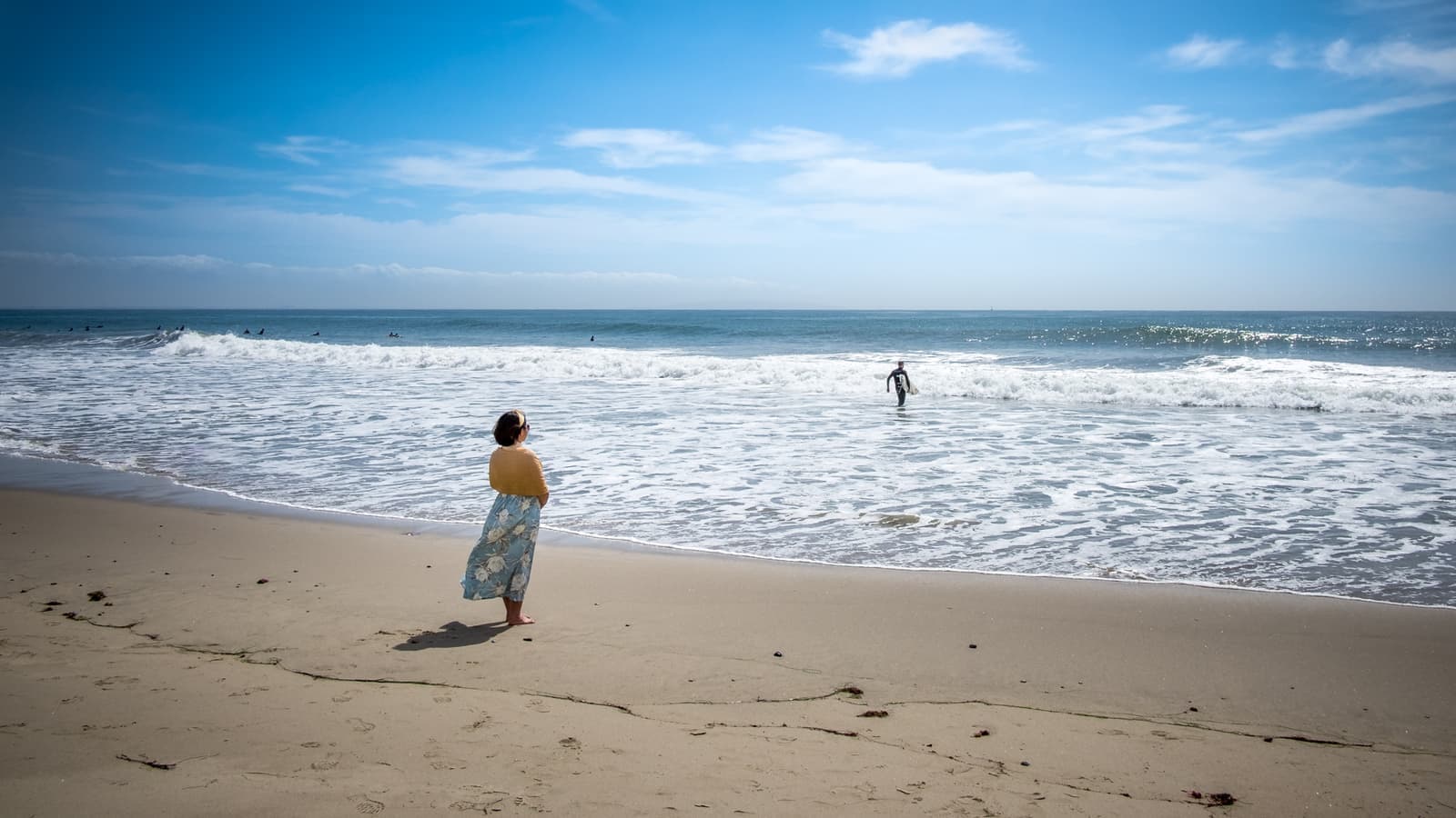 A woman in a long skirt stands on wet sand watching a surfer wade out of foamy waves under a bright blue sky
