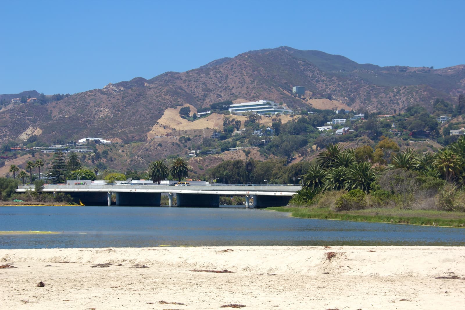 White sand before a calm blue lagoon, a concrete highway bridge with palms, and brown chaparral hills dotted with houses